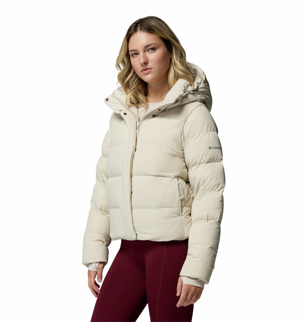 Une femme portant un blouson beige clair à capuche et un legging bordeaux se tient sur un fond blanc uni, regardant l'appareil photo avec assurance.