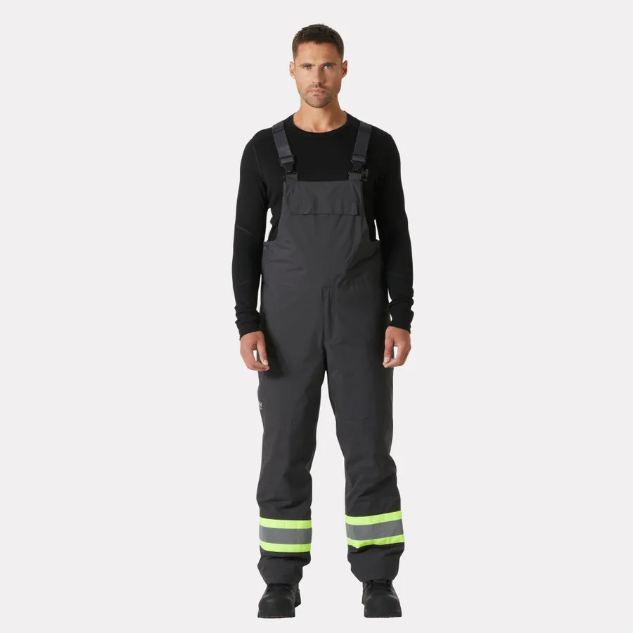 Un homme portant une salopette isotherme ALTA WINTER BIB CSA gris foncé, dotée de bandes réfléchissantes au niveau des jambes, par-dessus une chemise noire à manches longues.
