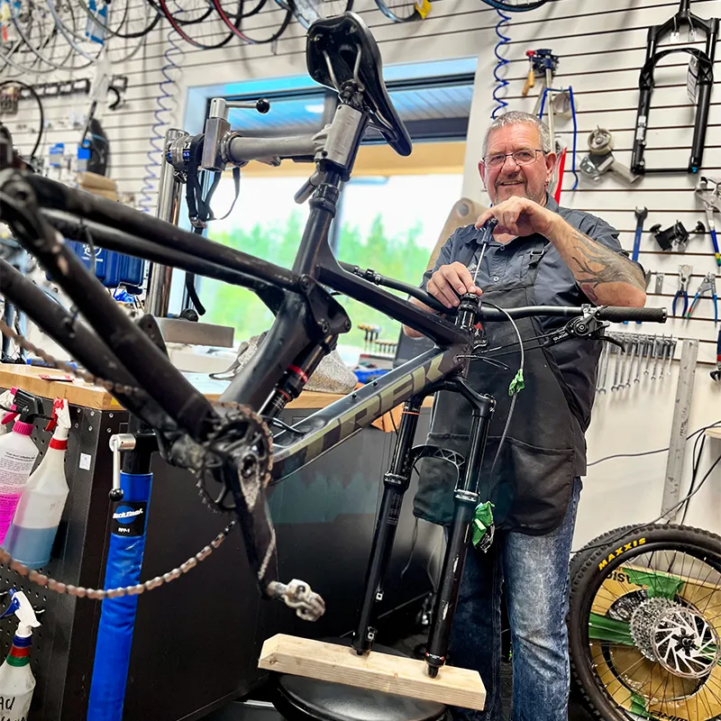 Un mécanicien cycliste souriant travaille sur un vélo noir dans un magasin de sport, entouré d'outils, de pièces de vélo et de roues suspendues au mur derrière lui.