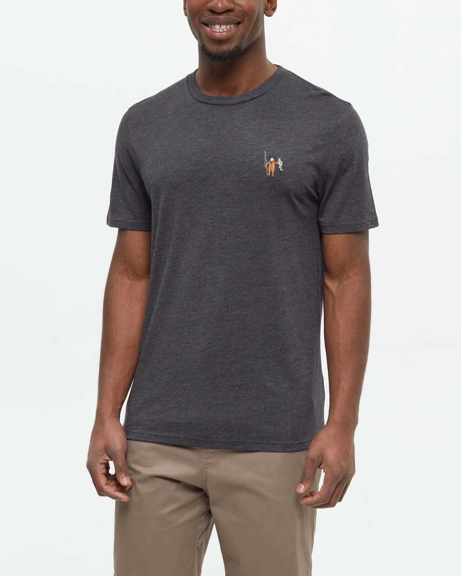 Un homme souriant portant un t-shirt à manches courtes gris foncé avec un petit motif brodé sur la poitrine et un pantalon marron clair se tient sur un fond blanc uni.