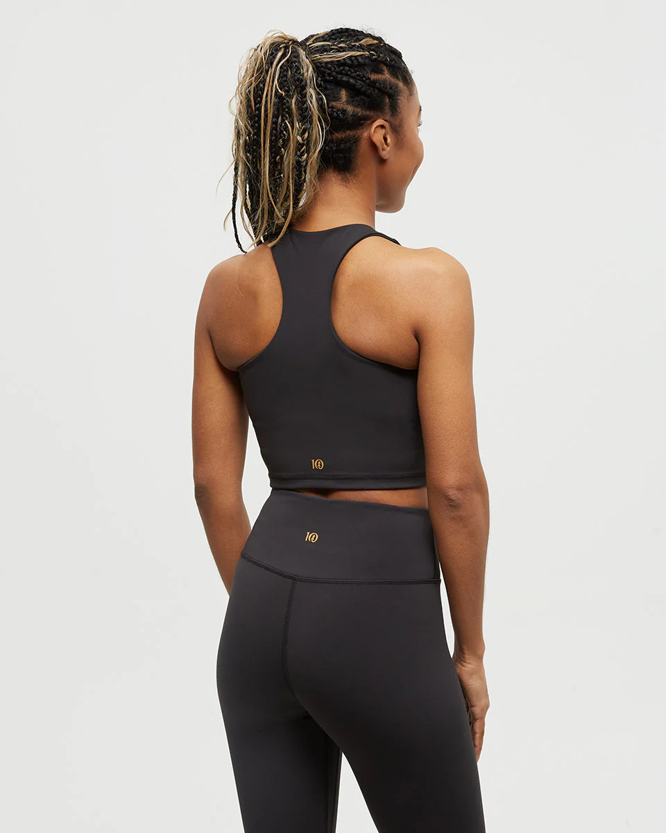 Une femme aux cheveux tressés porte un soutien-gorge de sport noir et un legging taille haute, debout, dos à la caméra sur un fond blanc uni.