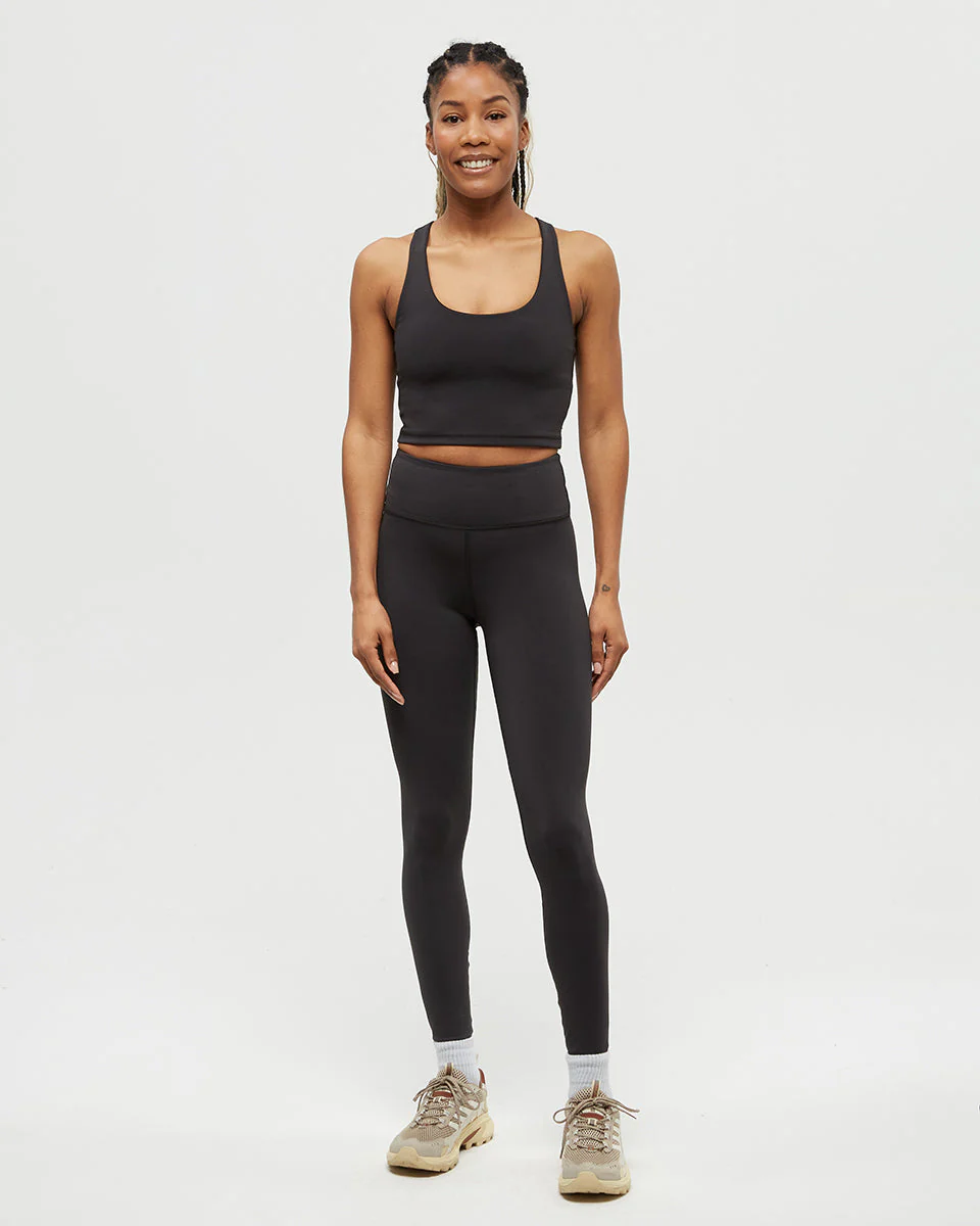 Une femme se tient debout, souriante, sur un fond blanc uni, portant un soutien-gorge de sport noir, des leggings noirs, des chaussettes blanches et des baskets beiges.