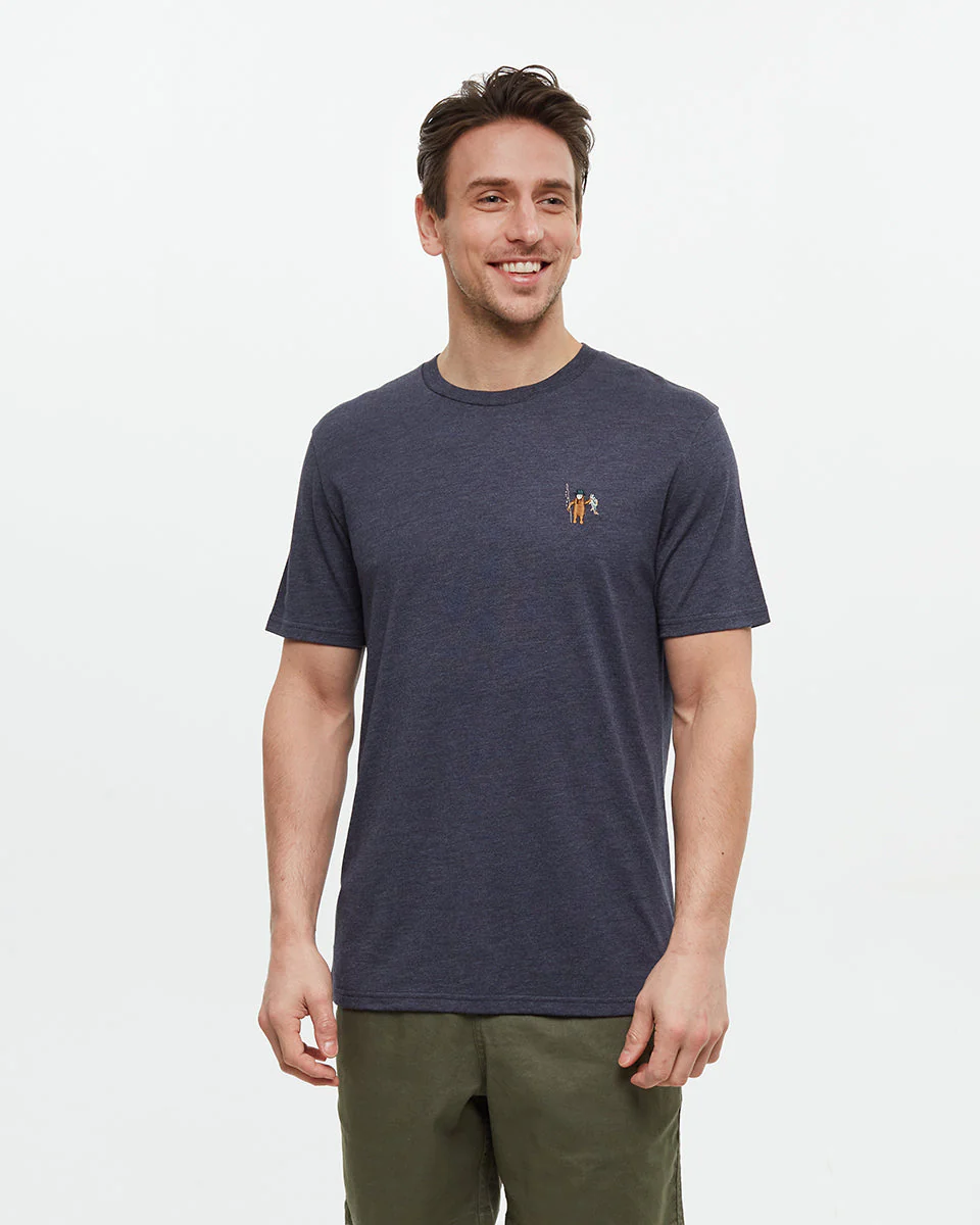 Un homme portant un t-shirt bleu foncé à manches courtes et un pantalon vert olive sourit sur un fond blanc uni. Le t-shirt arbore un petit motif brodé sur le côté gauche de la poitrine.