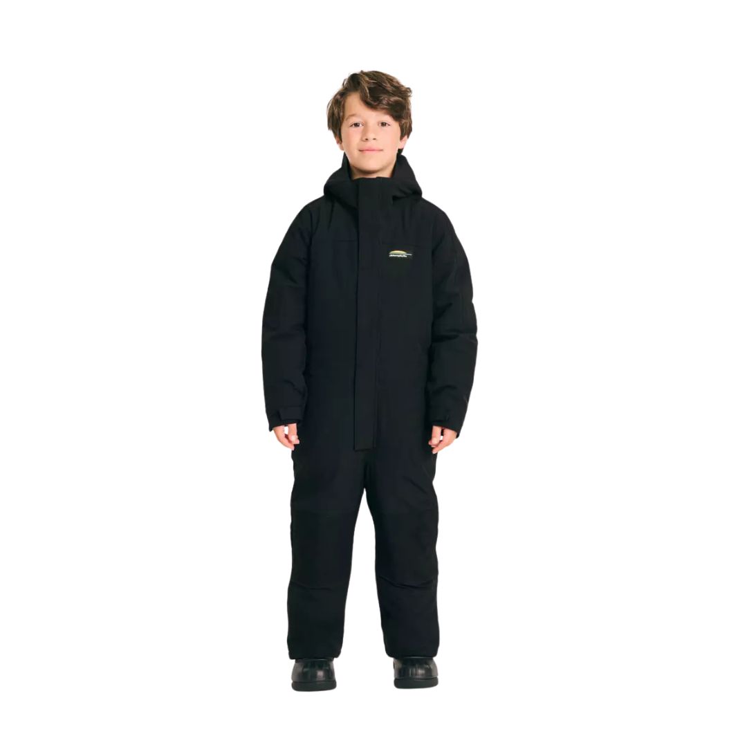Un jeune garçon portant un costume d'hiver noir à capuche et des bottes noires se tient debout, de face, sur un fond blanc uni.