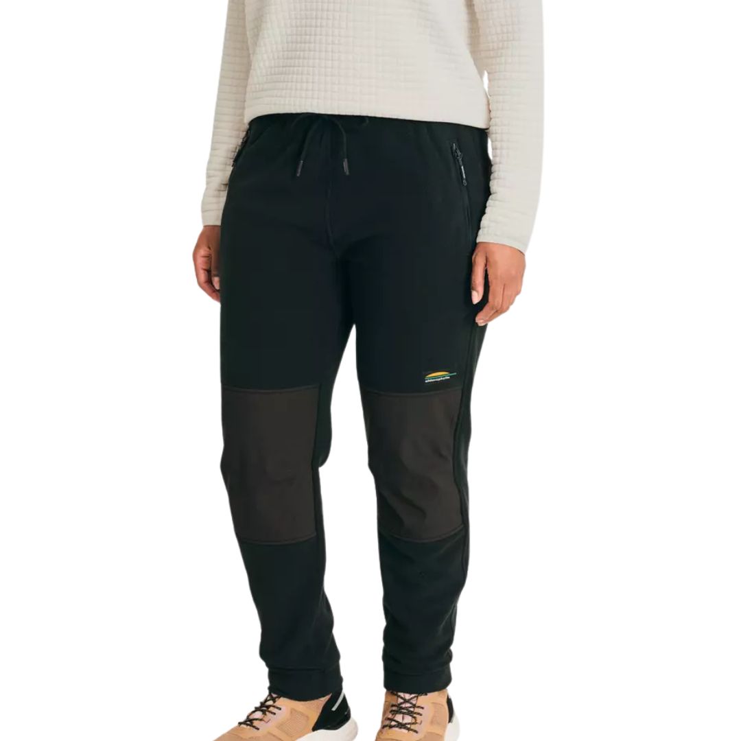 Une personne portant un pantalon noir de type jogger avec des renforts aux genoux, un haut blanc texturé à manches longues et des baskets beiges. Le pantalon a des poches zippées et un petit logo coloré sur la cuisse gauche.
