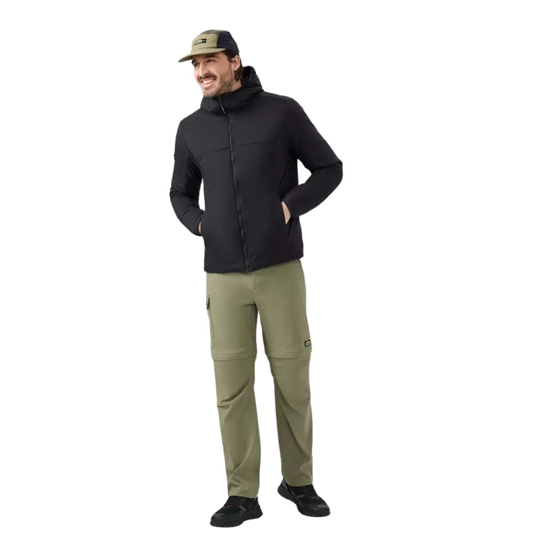 Un homme souriant, les mains dans les poches, vêtu d'une veste zippée noire, d'un pantalon vert olive, de chaussures noires et d'une casquette de baseball beige, isolé sur un fond blanc.