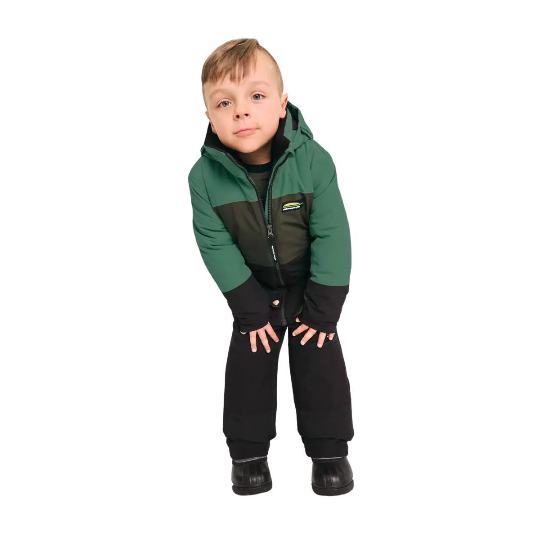 Un jeune garçon portant une veste d'hiver verte et noire et un pantalon de neige noir se penche en avant, les mains sur les genoux, en regardant l'appareil photo sur un fond blanc.