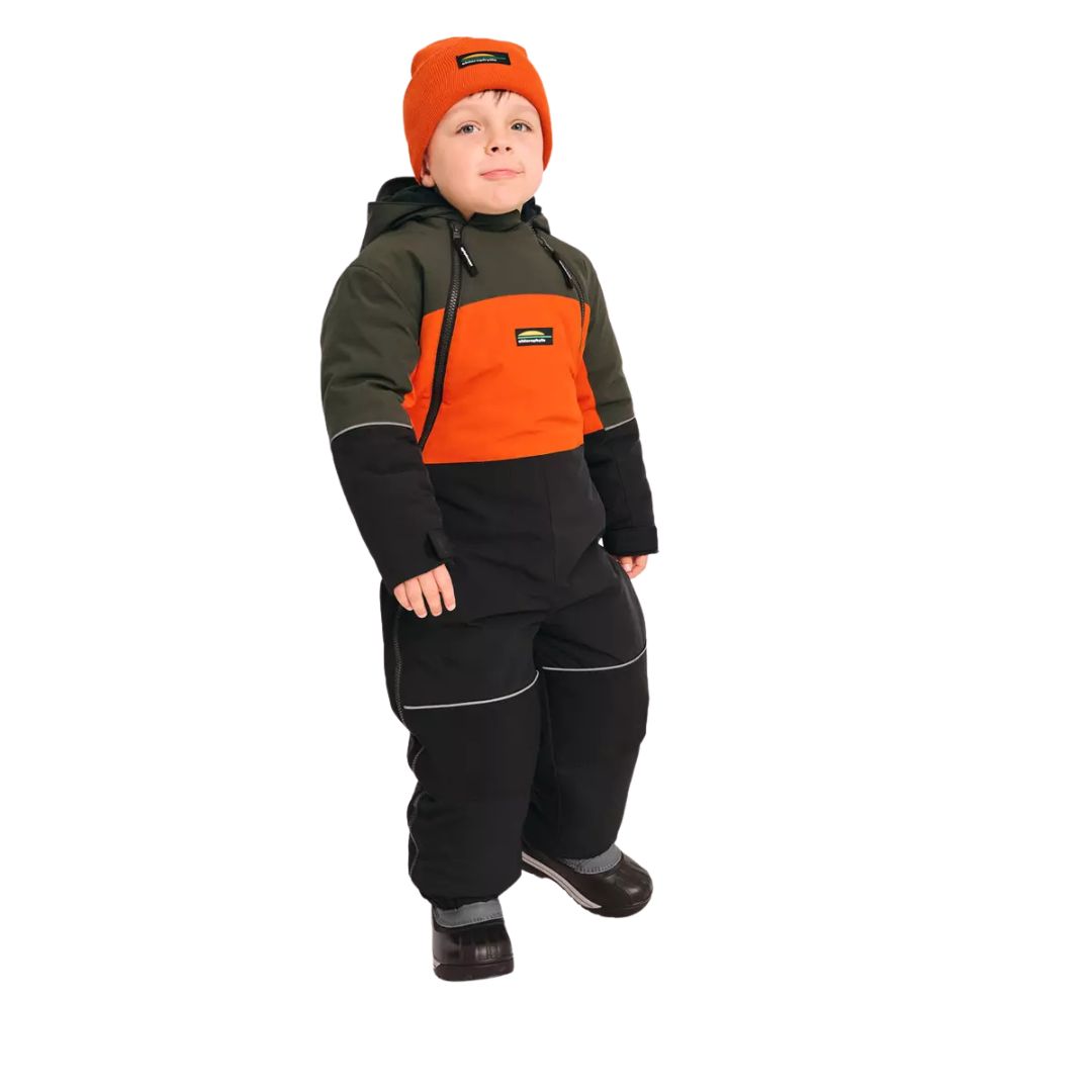 Un enfant portant l'HABIT DE NEIGE CHLOROPHYLLE CRICKET POUR ENFANT, un habit de neige vert, orange et noir, assorti d'un bonnet orange et de bottes noires, debout sur un fond blanc avec un léger sourire.