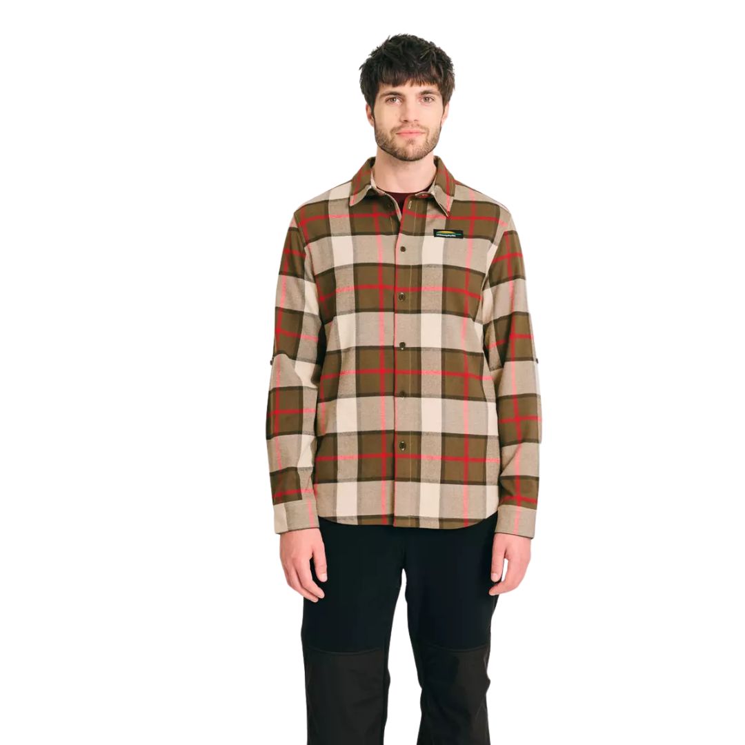Un homme se tient debout, de face, vêtu d'une chemise à boutons à carreaux beige, marron, vert et rouge et d'un pantalon noir, sur un fond blanc uni.