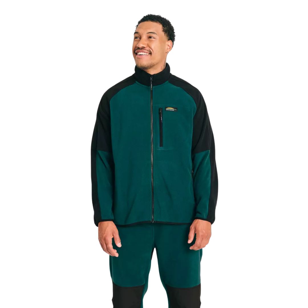 Un homme souriant, vêtu d'une veste polaire zippée sarcelle et noire avec une poche sur la poitrine et un pantalon sarcelle assorti, sur un fond blanc uni.