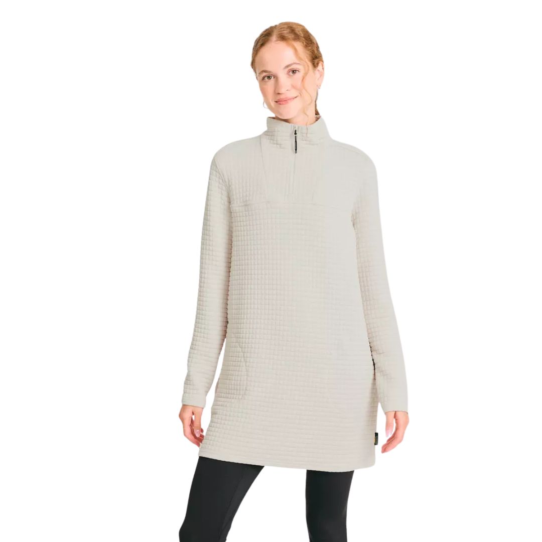Une femme vêtue d'une tunique gaufrée gris clair à manches longues, à col montant et à fermeture éclair sur le devant, associée à un legging noir, se tient sur un fond blanc uni, avec un léger sourire.