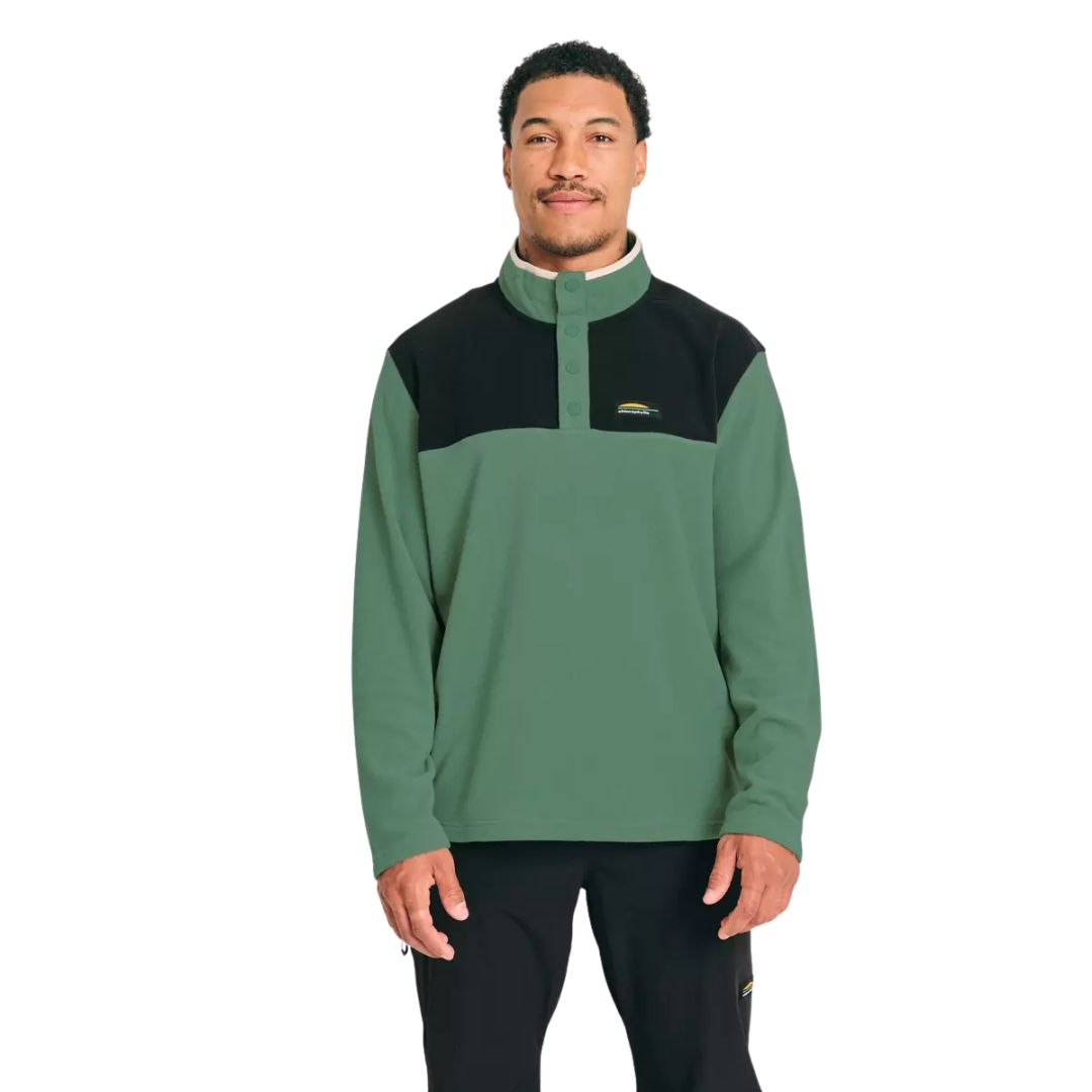 Un homme se tient debout, de face, vêtu d'un pull vert et noir à col boutonné et d'un pantalon noir. Il a les cheveux courts et une barbe taillée, et sourit légèrement sur un fond blanc uni.