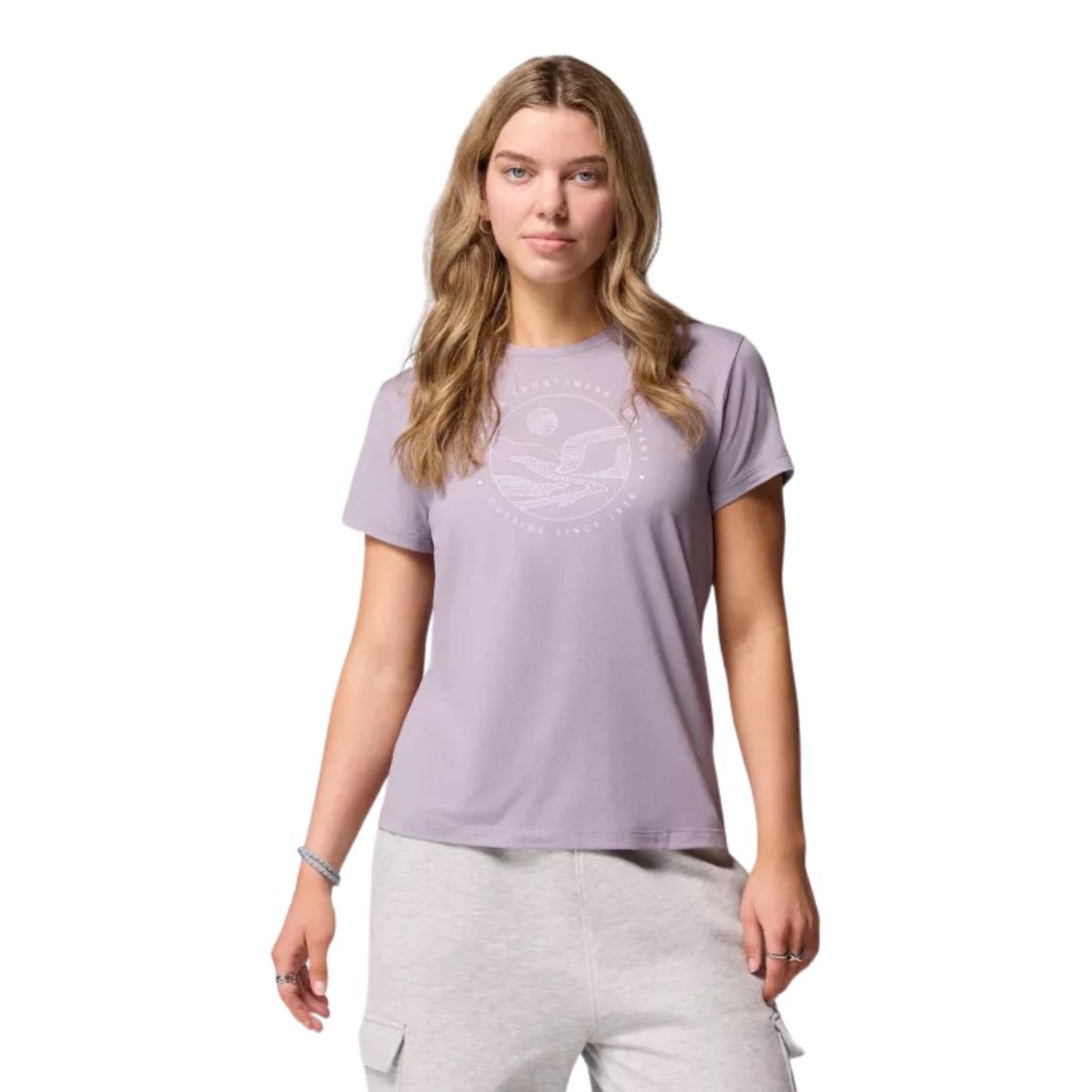 Une femme aux longs cheveux blonds ondulés porte un t-shirt violet clair avec un graphisme subtil et un pantalon cargo gris clair, sur un fond blanc uni.