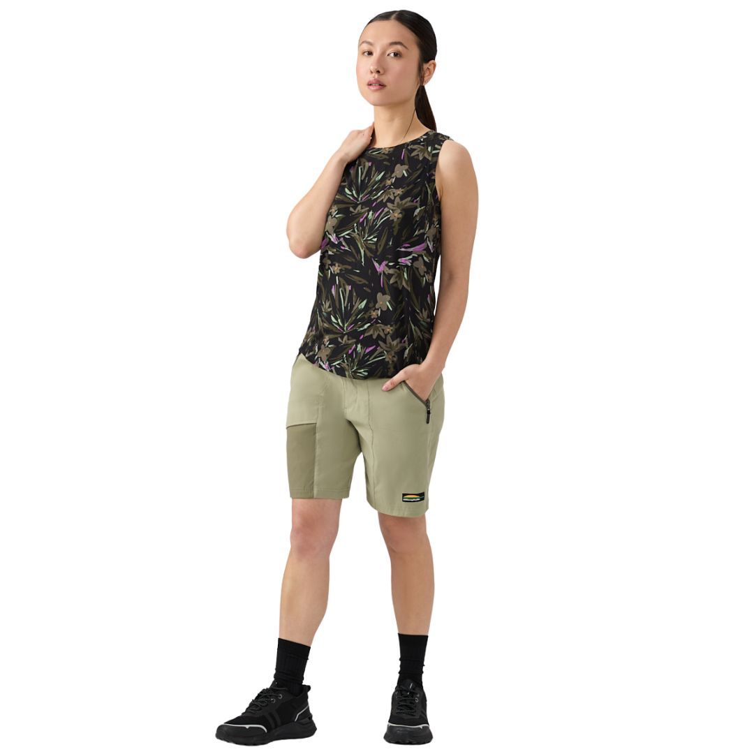 Une femme se tient debout, une main dans la poche de son short, portant un haut noir sans manches avec un imprimé feuillu, un short kaki clair, des chaussettes noires et des baskets noires sur un fond blanc uni.