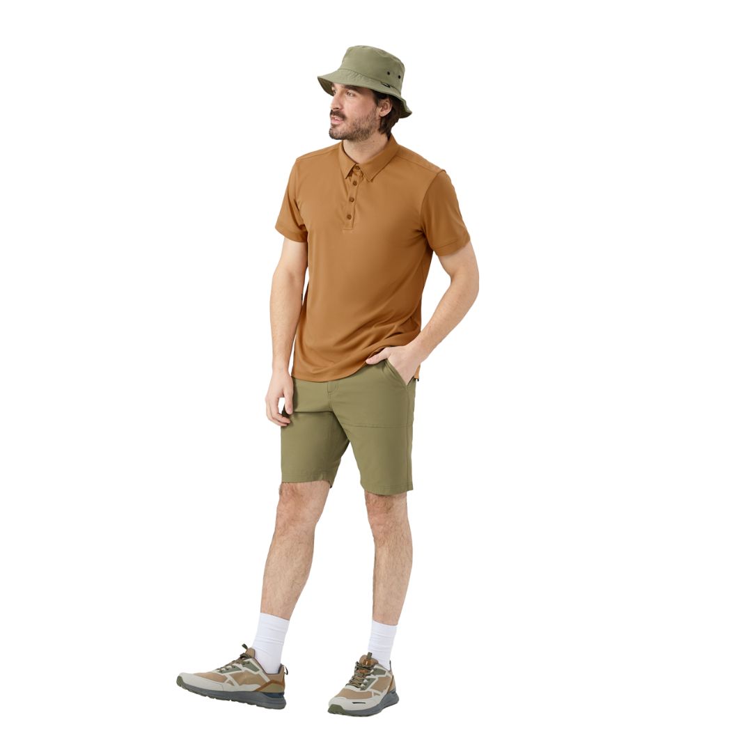 Un homme portant un chapeau seau vert, un polo marron, un short vert olive, des chaussettes blanches et des baskets marron se tient debout sur un fond blanc avec une main dans sa poche, regardant sur le côté.