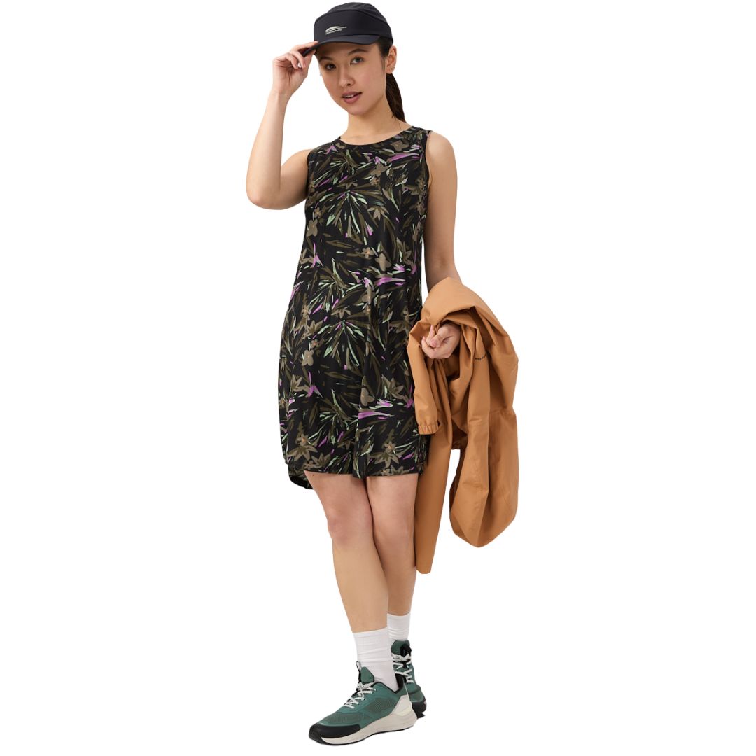 Une femme portant une robe à imprimé floral sans manches, des baskets vertes, des chaussettes blanches et une casquette noire tient une veste beige dans une main et touche sa casquette avec l'autre, debout sur un fond blanc.