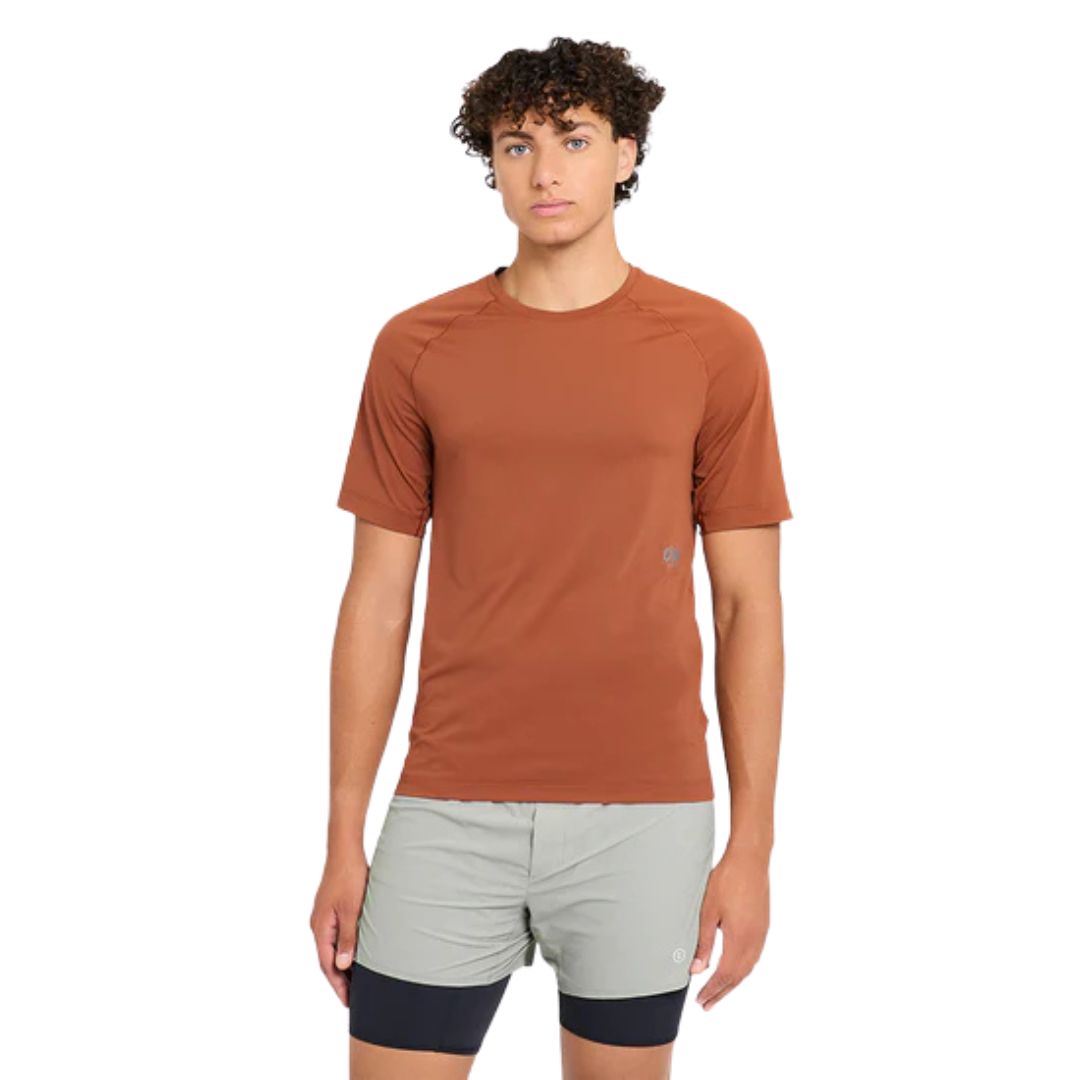 Une personne porte le T-SHIRT M DLY de couleur rouille avec un short gris clair doté d'une doublure de compression noire, debout, de face, sur un fond blanc uni.