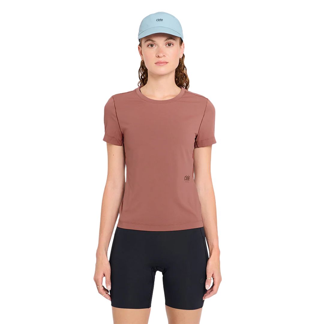 Une femme se tient de face sur un fond blanc uni, portant le T-SHIRT W DLY en mauve, un short de cyclisme noir et une casquette bleu clair.