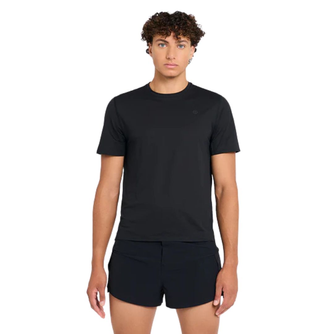 Une personne aux cheveux courts et bouclés portant le M FST TSHIRT en noir et un short noir se tient de face sur un fond blanc.