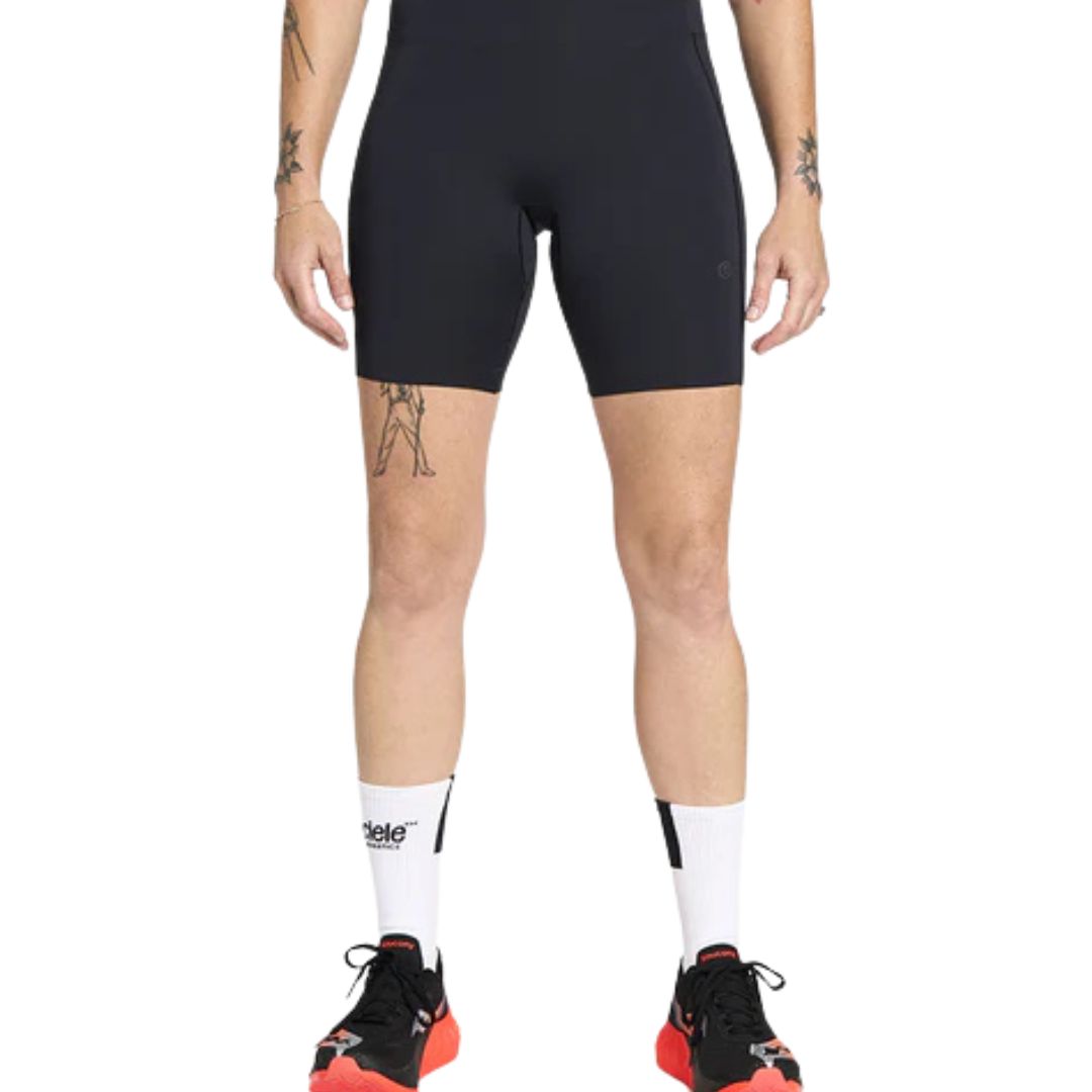 La personne porte un pantalon W FST TIGHT SHORT 7", des chaussettes blanches avec texte et des chaussures de sport noires et rouges. Des tatouages sont visibles sur les deux bras et la cuisse droite. L'arrière-plan est blanc.