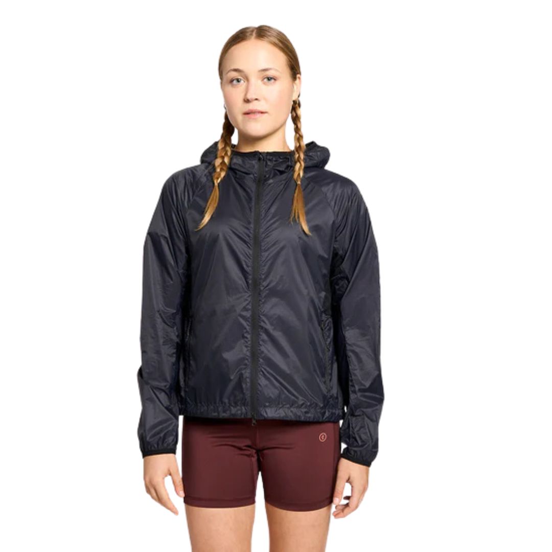 Une jeune femme aux cheveux tressés porte la VESTE DE PLUIE CIELE VNT POUR FEMME, une veste à capuche noire, associée à un short marron, sur un fond blanc uni.