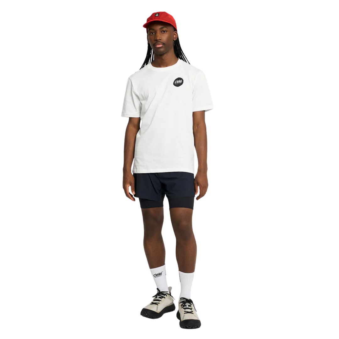 Une personne se tient debout, de face, sur un fond blanc uni, et porte l'encre U-ORTSHIRT (t-shirt blanc avec un petit logo noir), une casquette rouge, un short noir, des chaussettes blanches et des chaussures de sport blanches.