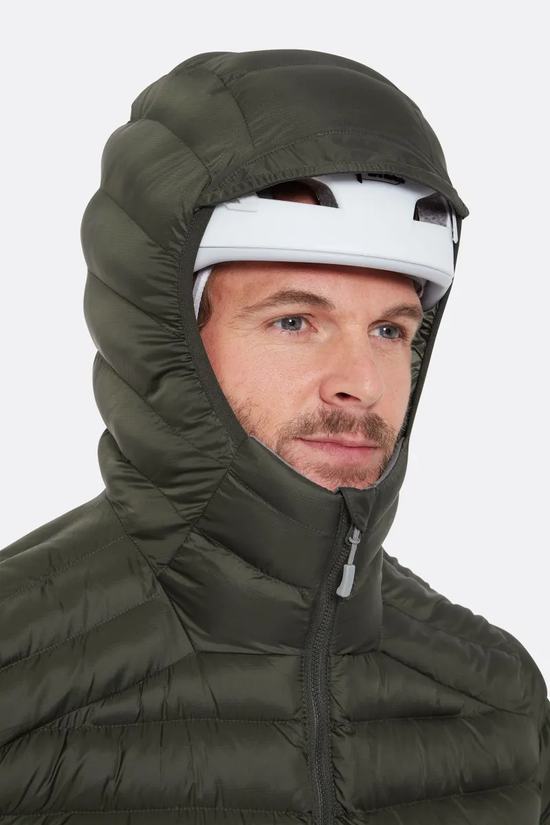 Une personne porte une doudoune vert foncé avec la capuche relevée, recouvrant un casque blanc. La veste est zippée et la personne regarde légèrement sur le côté sur un fond uni.
