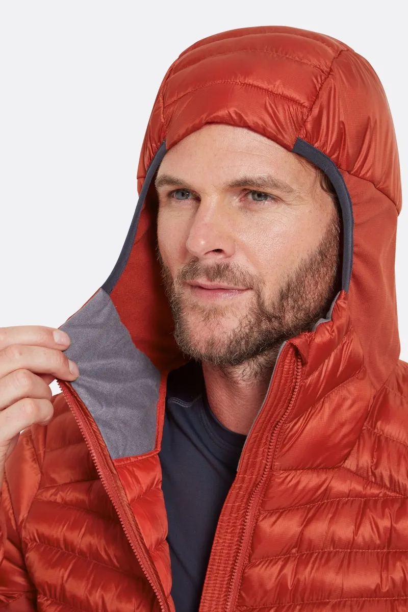 Une personne porte une doudoune à capuche orange. La veste est partiellement ouverte, révélant une doublure grise. La personne ajuste la capuche, elle a une barbe et les cheveux courts. L'arrière-plan est uni et de couleur claire.