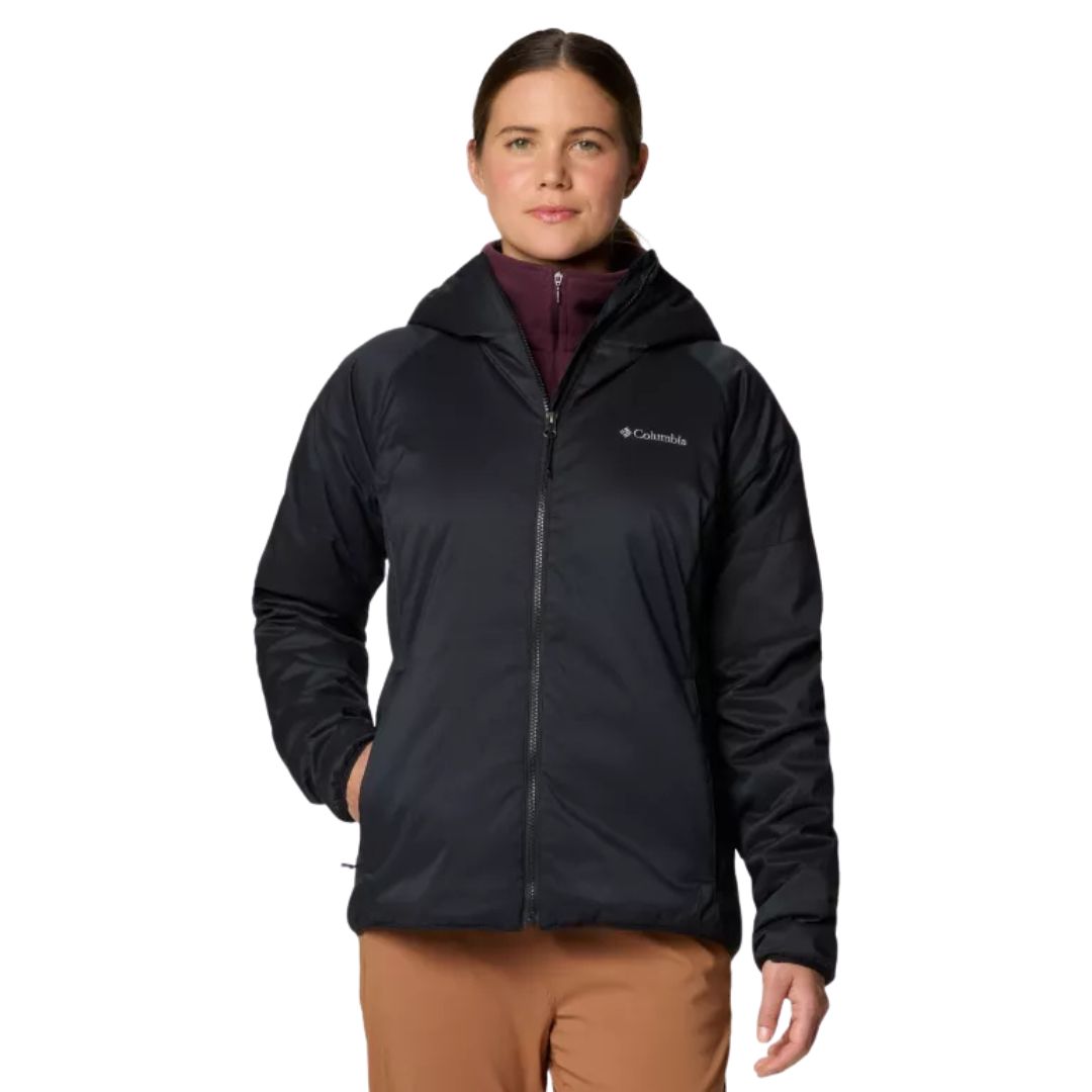 Une personne portant une veste à capuche Columbia noire sur un haut zippé marron et un pantalon beige se tient de face, une main dans la poche, sur un fond blanc uni.