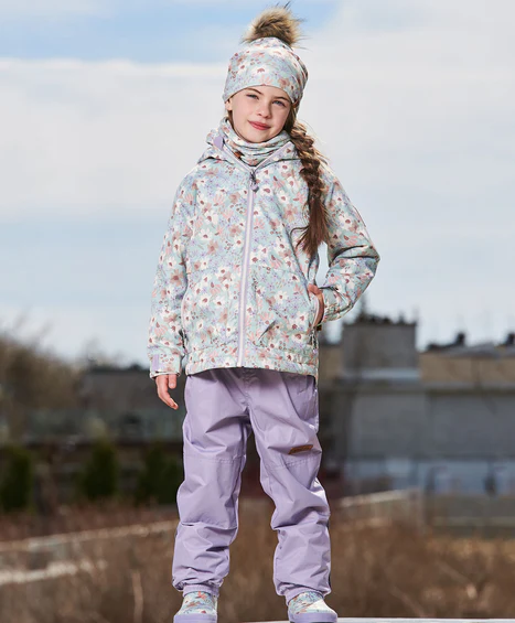 Une jeune fille se tient dehors par un jour nuageux, vêtue d'une veste à motifs floraux, d'un bonnet assorti à pompon et d'un pantalon violet. Ses longs cheveux sont tressés et elle semble se trouver sur un toit ou une terrasse surélevée.