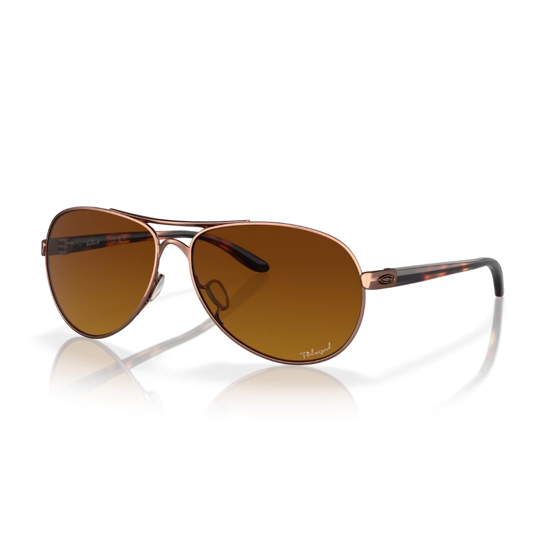 Une paire de lunettes de soleil aviateur Oakley Feedback Polished Gold avec des verres teintés en brun, une fine monture en métal et des branches en écailles noires et brunes sur fond blanc.