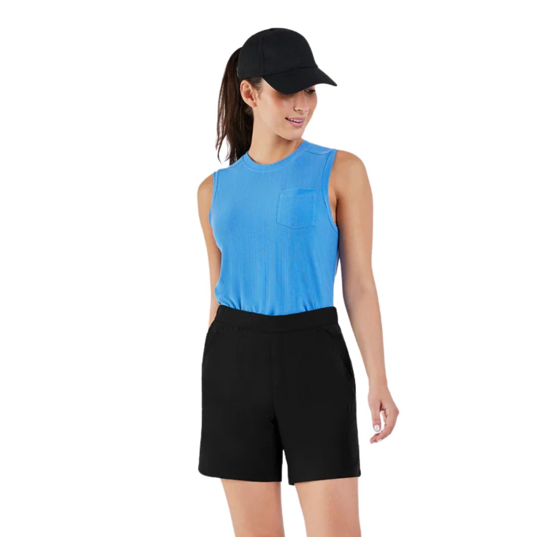 Une femme portant une casquette de baseball noire, un haut bleu sans manches avec une poche et un short noir se tient debout sur un fond blanc, regardant légèrement vers sa gauche.