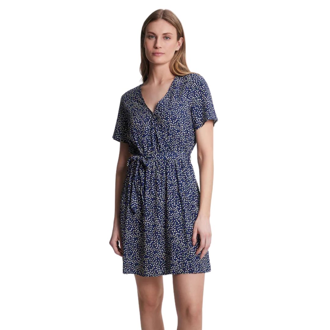 Une femme porte une robe bleu marine à manches courtes avec un motif à pois blancs et une taille nouée, se tenant debout sur un fond blanc uni.