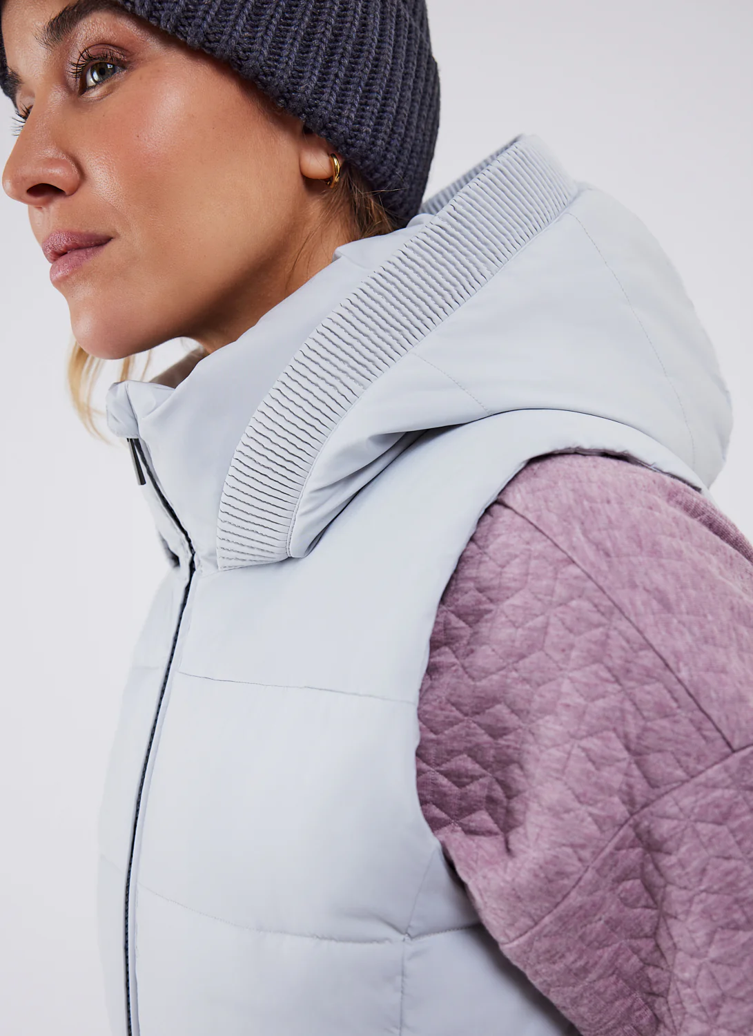 Une personne porte un bonnet gris, une veste matelassée violet clair et un gilet à capuche gris clair. L'image est un profil latéral en gros plan, mettant l'accent sur les vêtements d'hiver confortables et superposés.