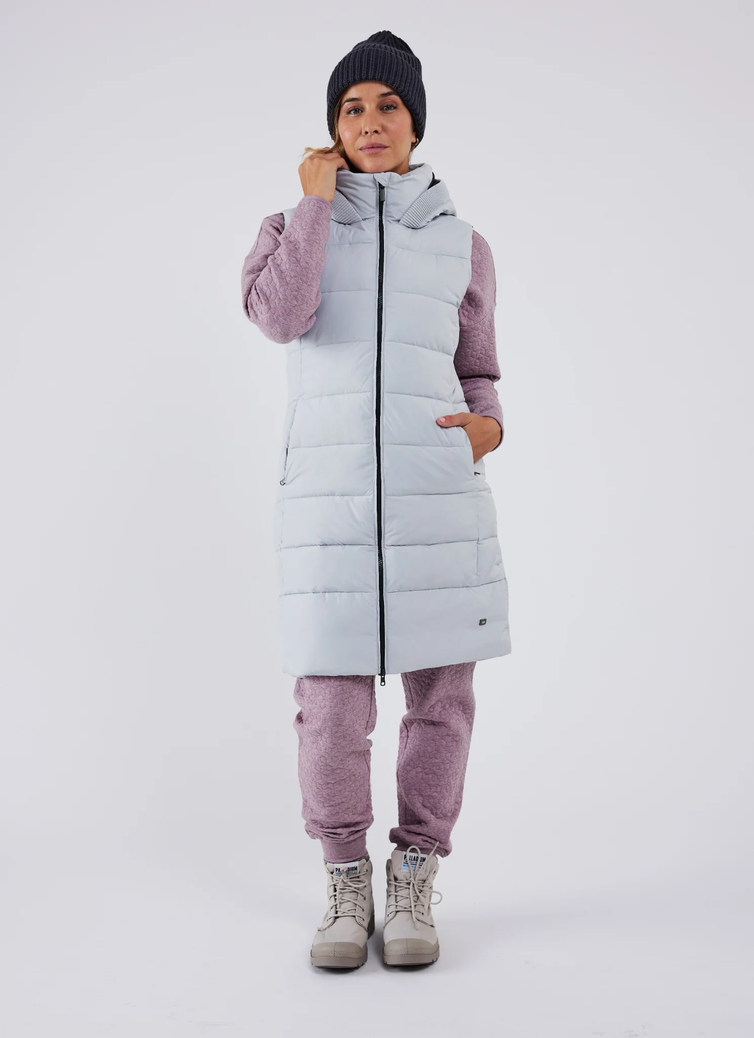Une personne portant un long gilet matelassé de couleur claire sur un sweat-shirt et un pantalon mauves, associés à des bottes beiges et un bonnet foncé, se tient debout sur un fond uni.