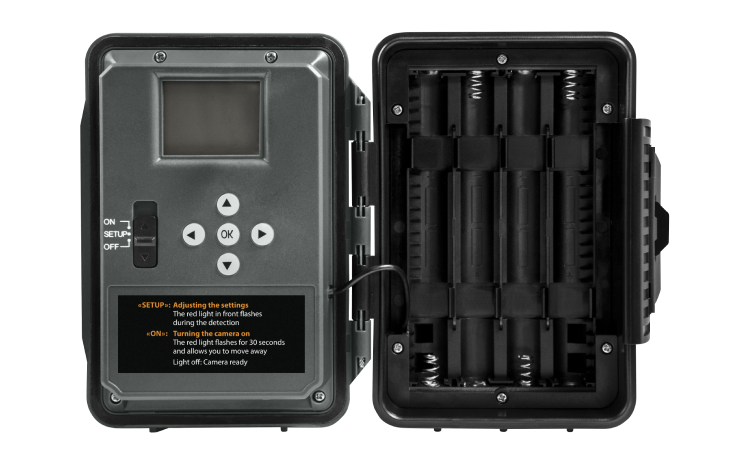 Une caméra de surveillance rectangulaire noire avec son couvercle avant ouvert, révélant un petit écran, des boutons de commande et un compartiment à piles. Le côté gauche contient des boutons étiquetés ON, SETUP et OFF, tandis que le côté droit contient des emplacements pour les piles.
