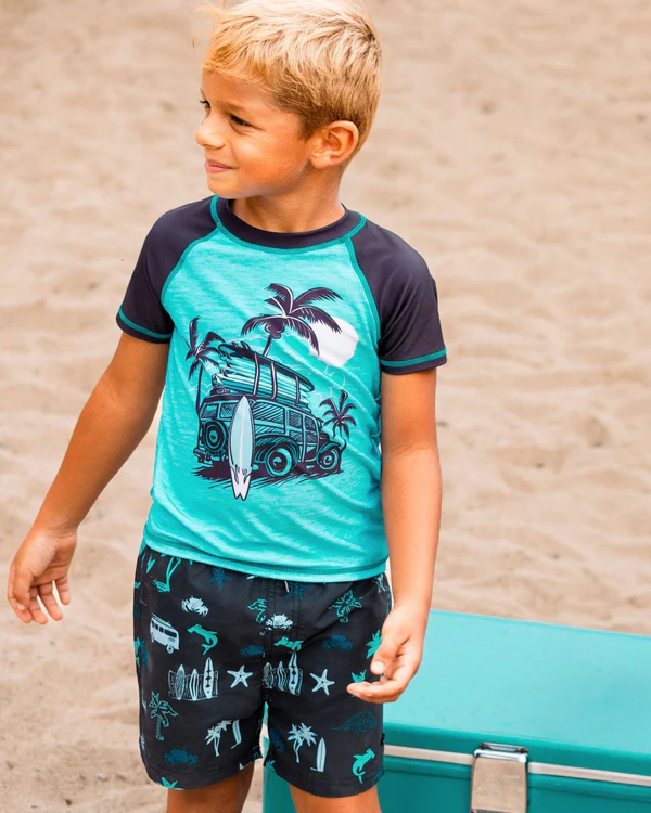 Un jeune garçon aux cheveux blonds courts se tient debout sur le sable, vêtu d'un rashguard turquoise et bleu marine orné d'un motif de van, de planche de surf et de palmier. Il porte un short assorti aux imprimés de plage et se tient près d'une glacière bleu sarcelle.