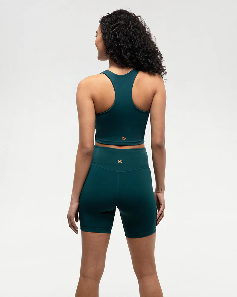 Une femme aux cheveux bouclés se tient debout, le dos tourné, vêtue d'un soutien-gorge de sport dos nageur bleu sarcelle foncé et d'un short cycliste taille haute assorti. Le fond est uni et gris clair.