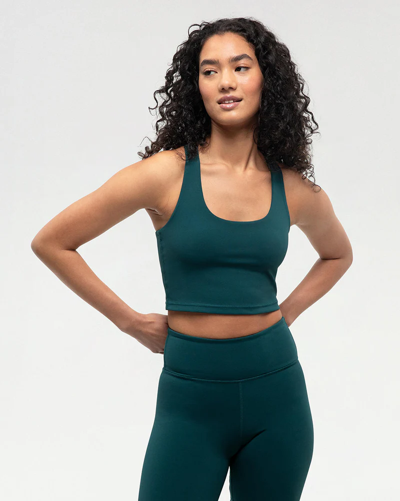 Une femme aux cheveux longs et bouclés portant un soutien-gorge de sport vert foncé et un legging taille haute assorti se tient debout, les mains sur les hanches, regardant sur le côté et souriant légèrement sur un fond clair uni.