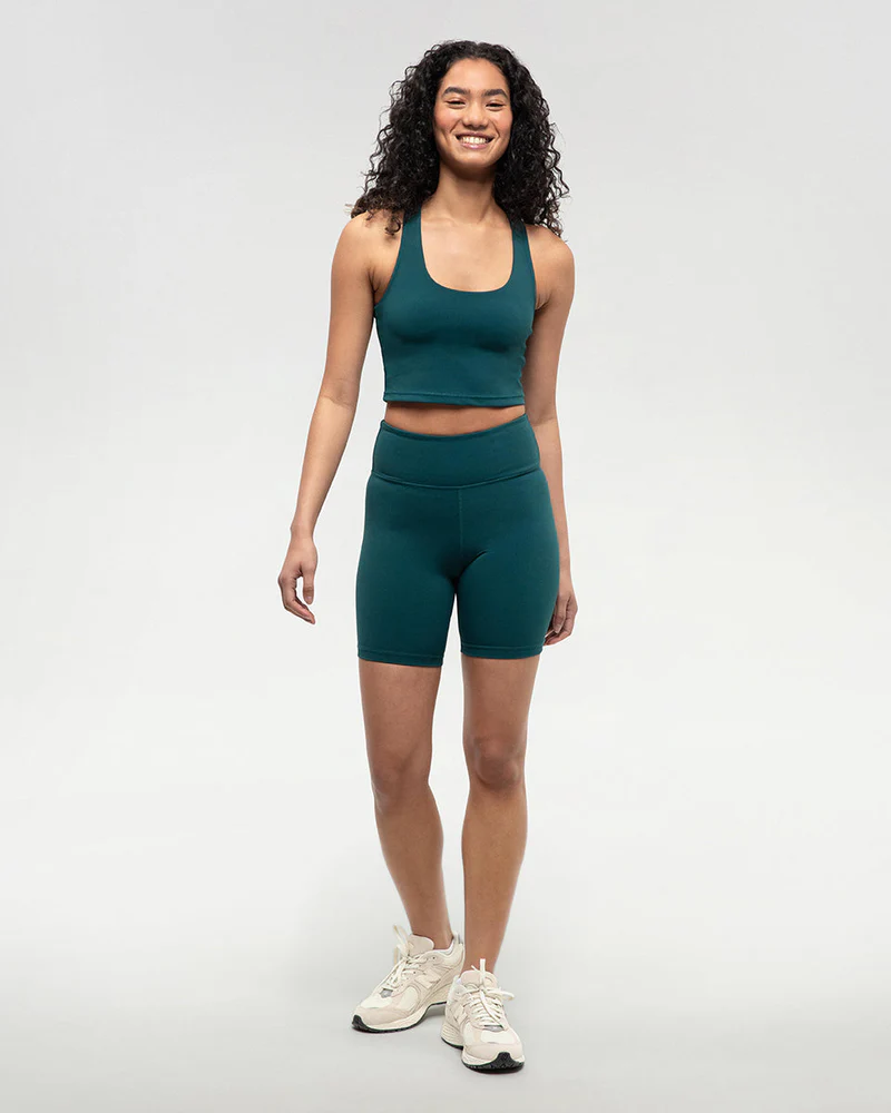 Une femme aux cheveux bouclés sourit et se tient debout sur un fond clair et uni. Elle porte un haut court de sport bleu sarcelle assorti, un short cycliste taille haute et des baskets blanches.
