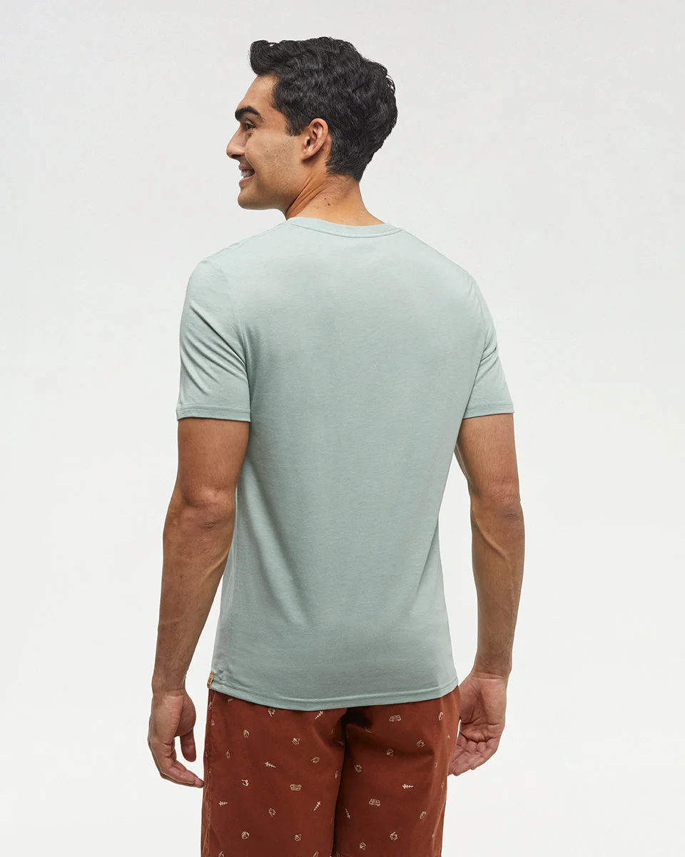 Un homme est debout, dos à la caméra, vêtu d'un t-shirt vert clair à manches courtes et d'un short marron à petits motifs. Il sourit, et l'arrière-plan est blanc uni.
