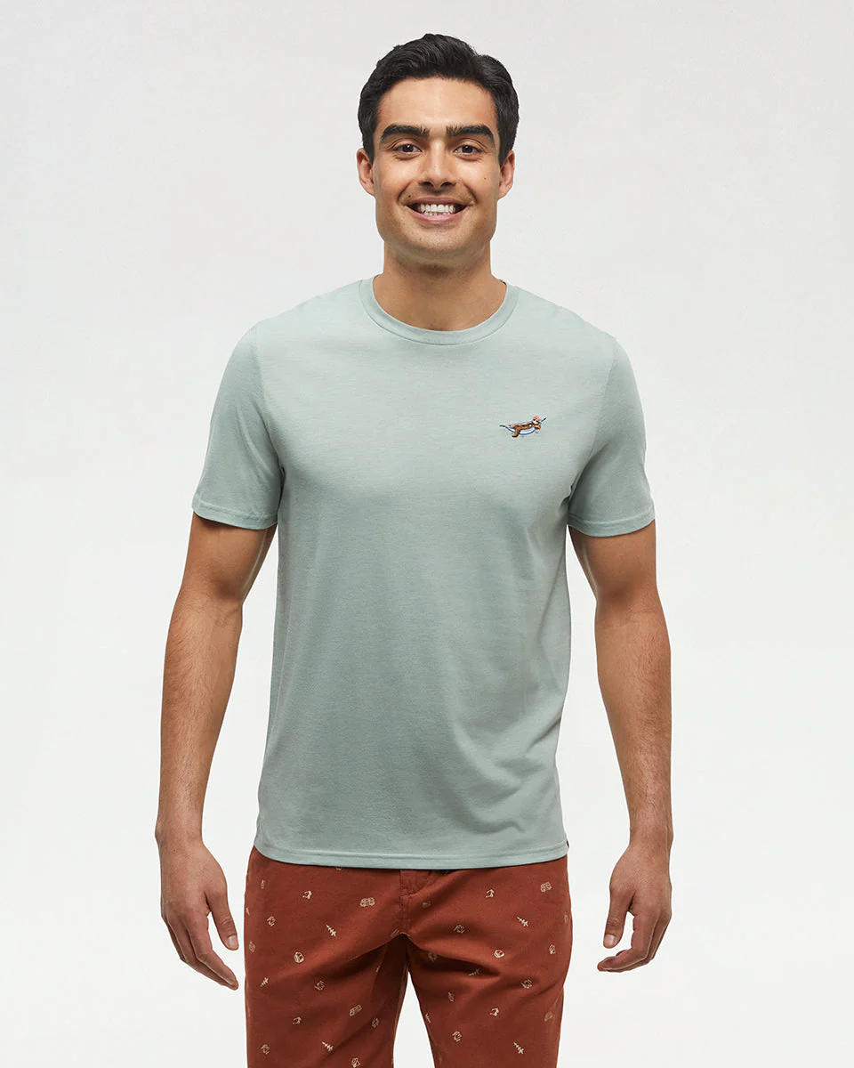 Un homme souriant se tient debout face vers l'avant, portant un t-shirt vert clair à manches courtes avec un petit motif brodé sur la poitrine et un pantalon couleur rouille avec un motif subtil, sur un fond clair uni.