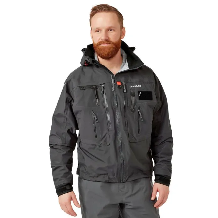 Un homme à la barbe rousse porte l'ALTA JACKET GRAPHITE 2017 - S, une veste d'extérieur gris foncé avec de multiples poches zippées et une capuche, associée à un pantalon assorti, sur fond blanc.