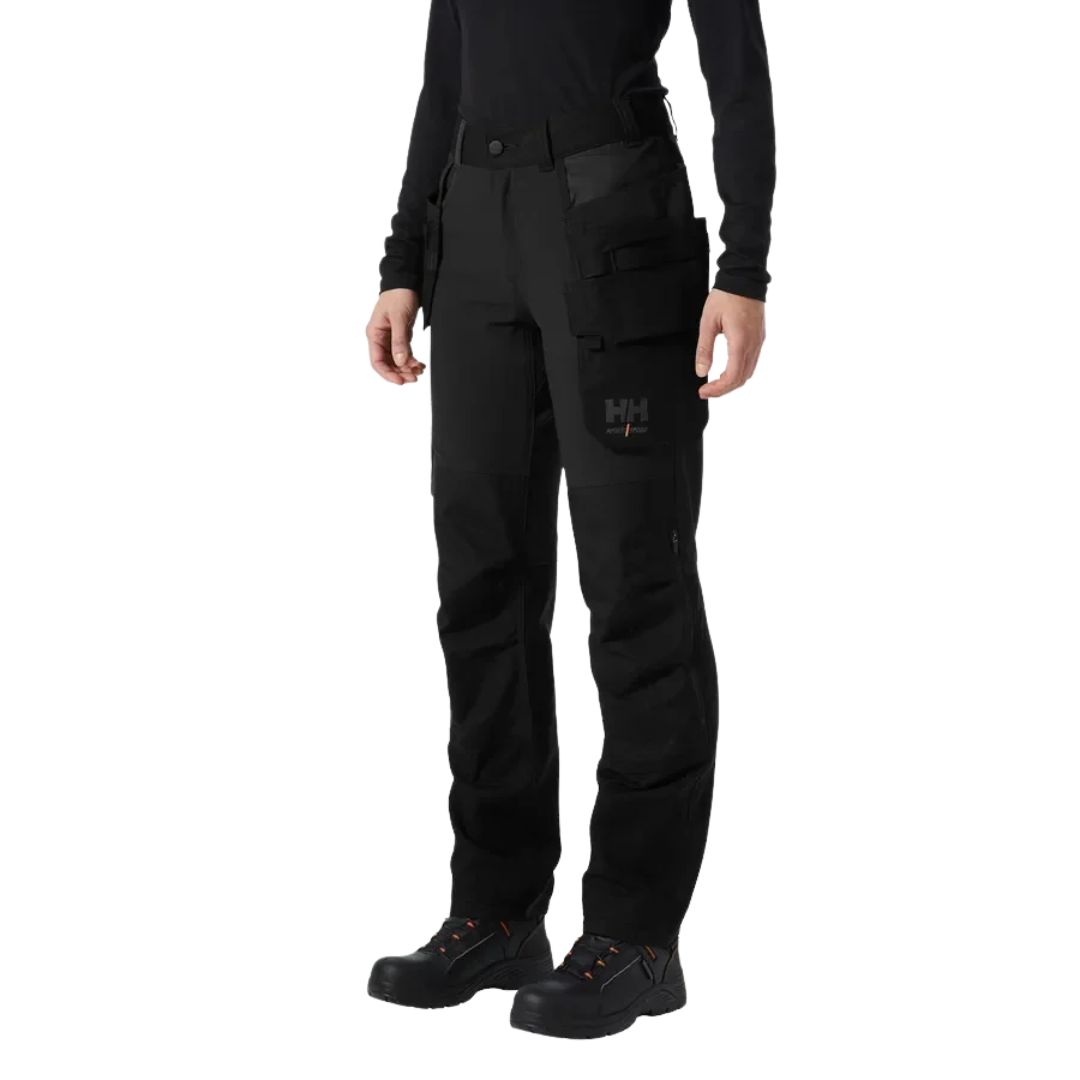 Pantalon de chantier noir HELLY HANSEN WORKWEAR LUNA 4X POUR FEMME, doté de poches fonctionnelles et d'un logo sur la cuisse, porté avec des bottes de travail noires.