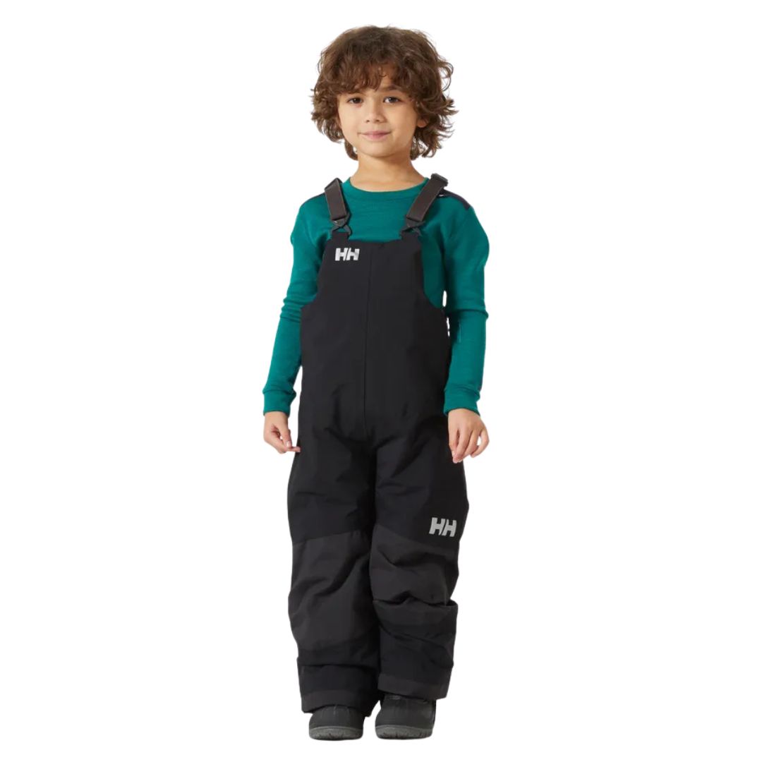 Un jeune enfant aux cheveux bruns bouclés porte une chemise à manches longues sarcelle et un pantalon de neige à bavette Helly Hansen noir. Il se tient debout et sourit sur un fond blanc uni.