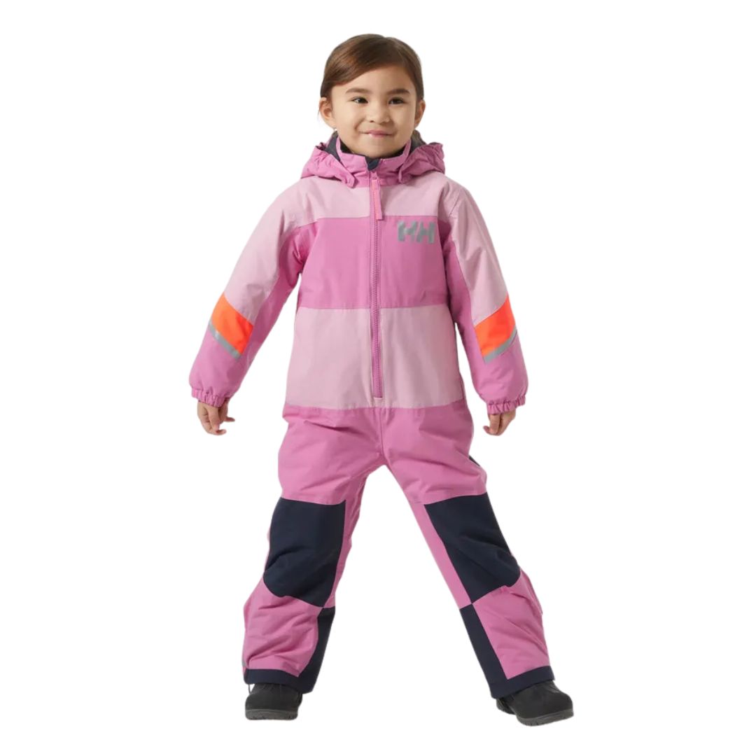 Un jeune enfant portant un habit de neige à capuchon rose et violet avec des bandes orange sur les manches et des taches noires sur les jambes, debout et souriant sur un fond blanc.