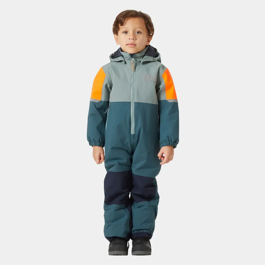 Un jeune enfant aux cheveux bruns courts se tient debout, tourné vers l'avant, vêtu d'une combinaison de neige aux couleurs contrastées avec des nuances de vert, de bleu sarcelle et de bleu marine, et des accents orange sur les épaules. Il porte également des bottes d'hiver noires. L'arrière-plan est blanc uni.