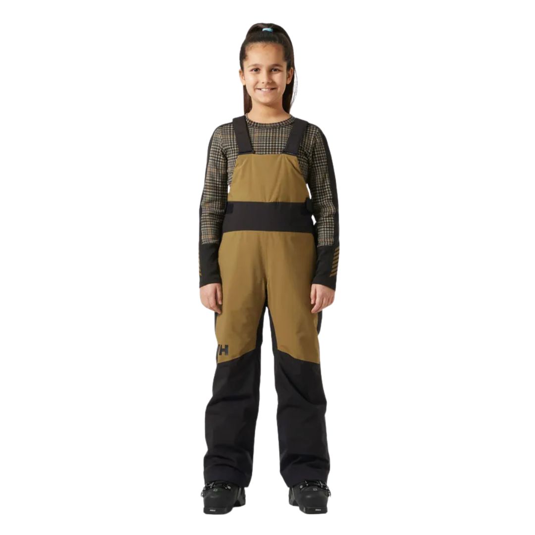 Une jeune fille souriante portant un habit de neige à bavette marron et noir sur une chemise à manches longues à motifs et des bottes de neige noires, debout sur un fond blanc uni.