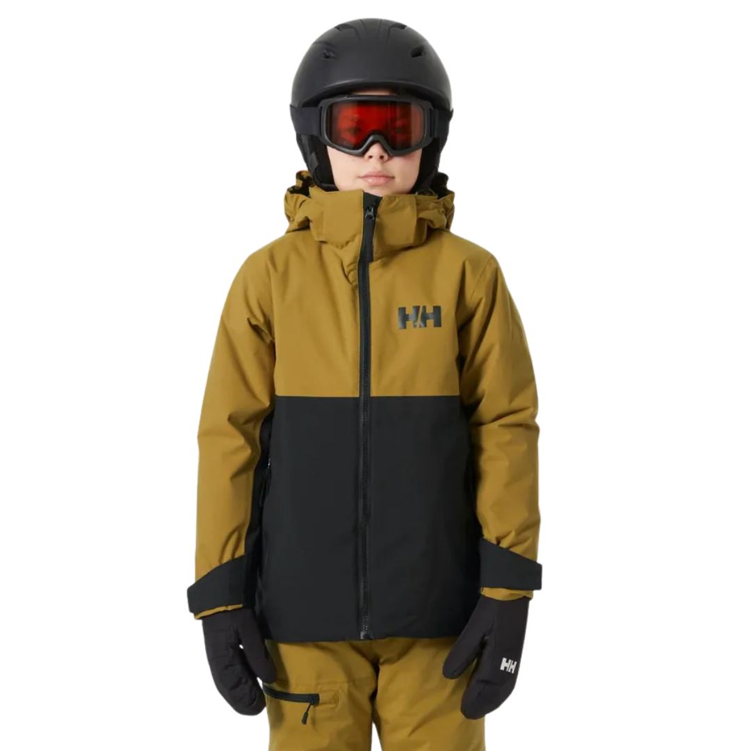 Un enfant portant un casque noir, des lunettes de ski rouges, une veste d'hiver Helly Hansen marron et noire, un pantalon marron assorti et des gants noirs se tient de face sur un fond blanc.