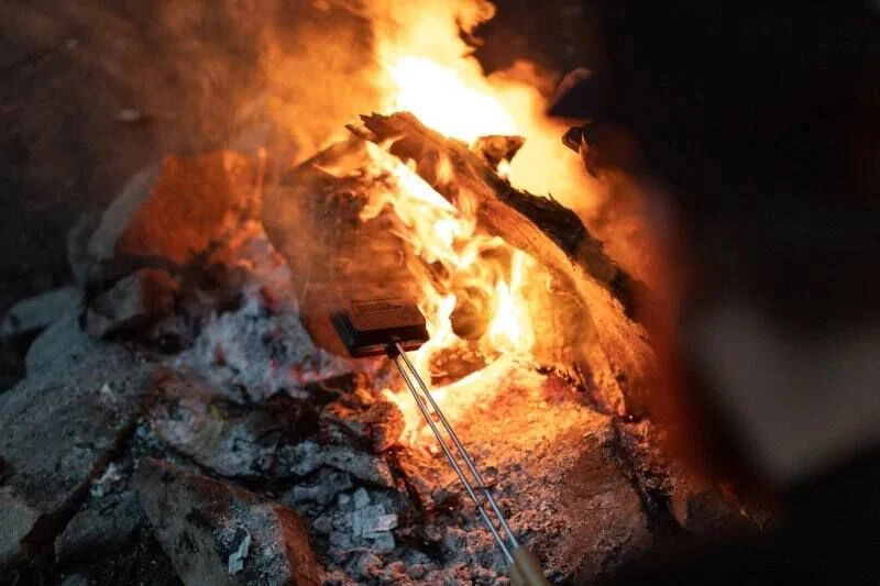 Un feu de camp brûle vivement, avec des flammes orange et des braises incandescentes. Une personne tient un panier à griller au-dessus du feu, en train de rôtir des aliments. La scène est entourée de rochers, ce qui donne une impression d'extérieur rustique.